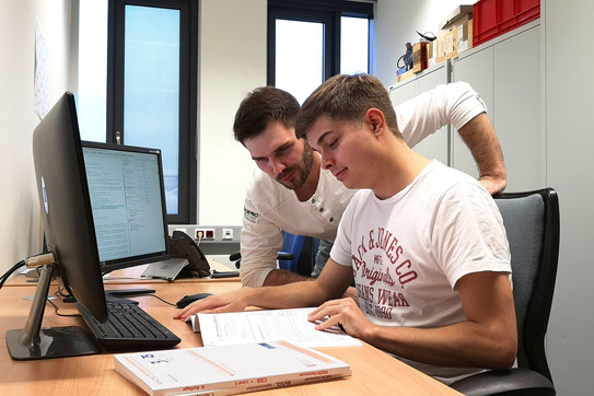 Two employees at a desk with two screens. They look into a book together.