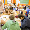 Mehrere Kinder sitzen an Tischgruppen in einem Klassenraum und lesen in Schulbüchern. Eine Lehrerin steht neben zwei Kindern und schaut mit ihnen in ein Buch.