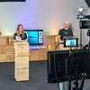 A woman and a man stand in front of two boxes and moderate an event. A camera is filming them.