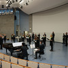 A choir including a conductor stands in an auditorium and sings, some choir members play an instrument.