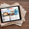 A tablet with a news website lies on a stack of newspapers