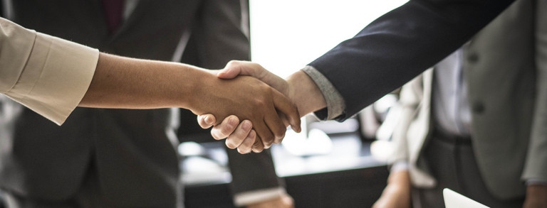 Two people shake hands. The handshake is shown in detail, in the background are two people in suits.