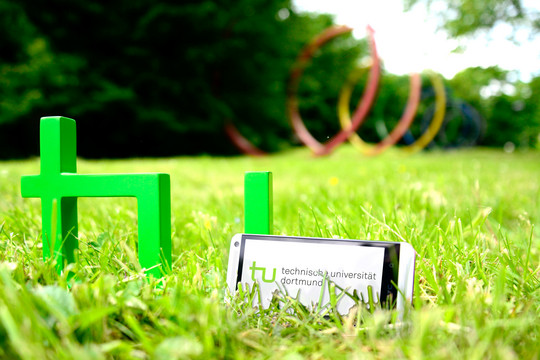 Foto eines Smartphones, das neben dem TU-Logo im Gras steht, im Hintergrund sind die Spektralringe zu sehen
