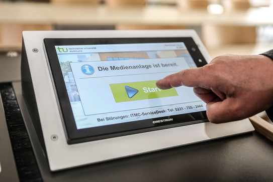 Eine Person tippt mit einem Finger auf einem Bildschirm am Hörsaalpult.